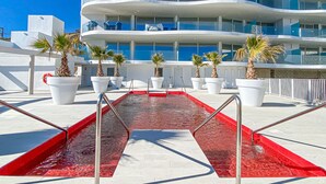 Un edificio moderno con varios balcones, una piscina con barandillas rojas de seguridad y palmeras en macetas.