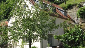 A white building with a red-tiled roof, surrounded by greenery and a stone wall. - Holiday Apartment Garden Paradise (Urbar)