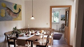 A dining area with a wooden table, chairs, and a bench. There is a painting on the wall, a hanging light fixture, and a view of the outdoors through a window. - Holiday Apartment Garden Paradise (Urbar)