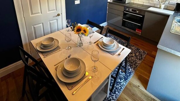 A dining area with a white table set for four, a clock on the wall, and a kitchen in the background. - Historic Old Town Home with Views and free parking (Kingston upon Hull)