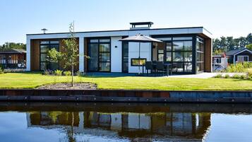 Een modern huis met twee verdiepingen, grote ramen en een plat dak, gelegen naast een rustig waterlichaam.