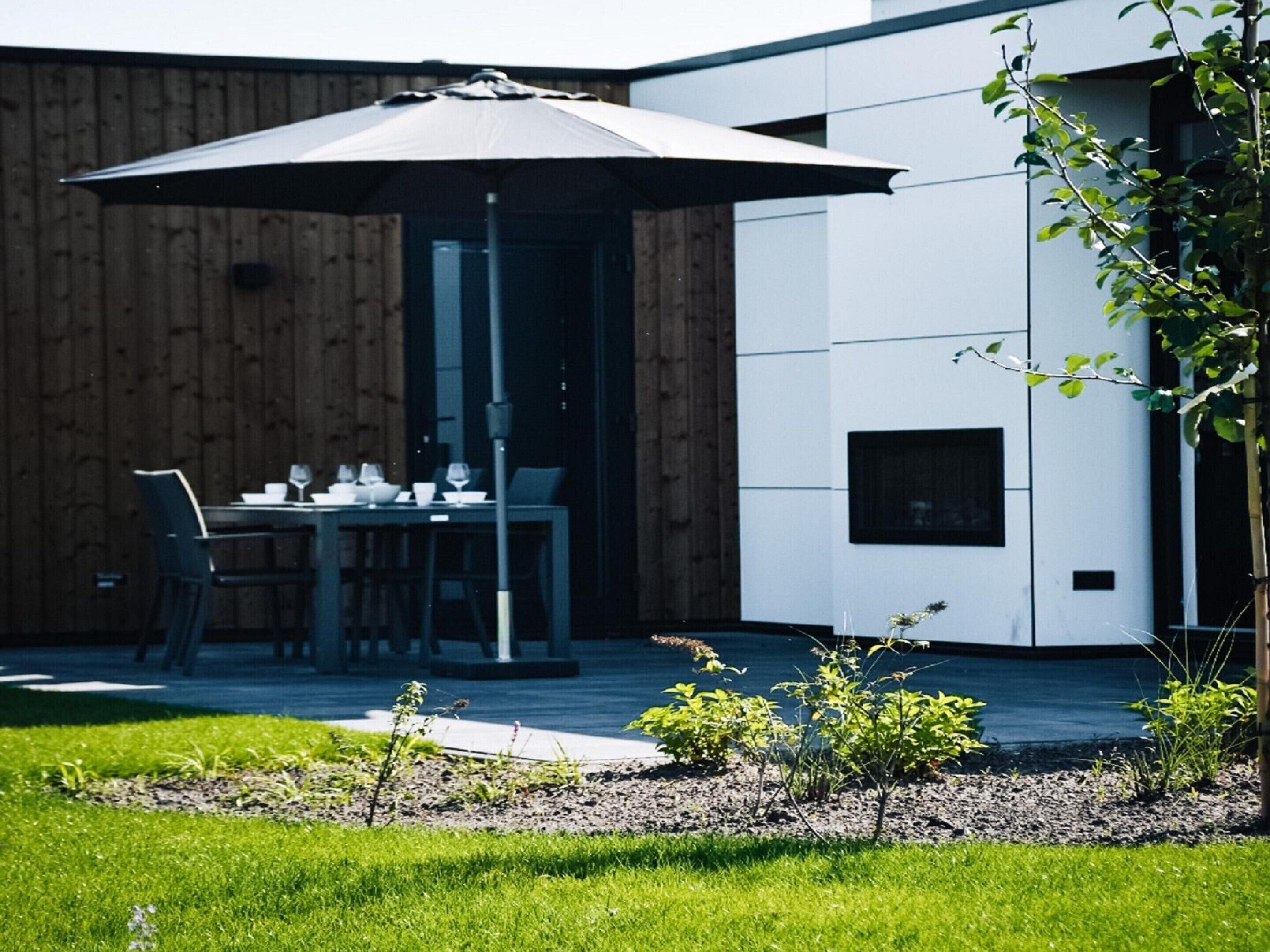 Een terras met een tafel en stoelen onder een grote parasol, naast een gebouw met een modern design en een open haard.