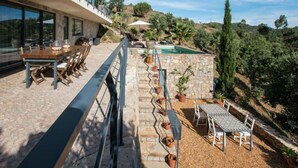 A modern outdoor dining area with a stone wall and potted plants, overlooking a pool and lush greenery. - Holiday Home Casa L (Sao Bras de Alportel)