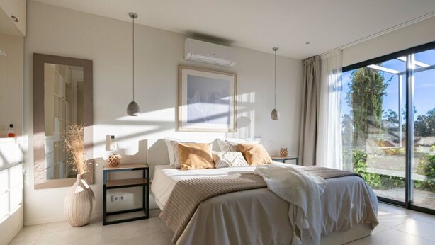 A modern bedroom with a large bed, a bedside table, a mirror, and a window with a view of greenery. - Holiday Home Casa L (Sao Bras de Alportel)