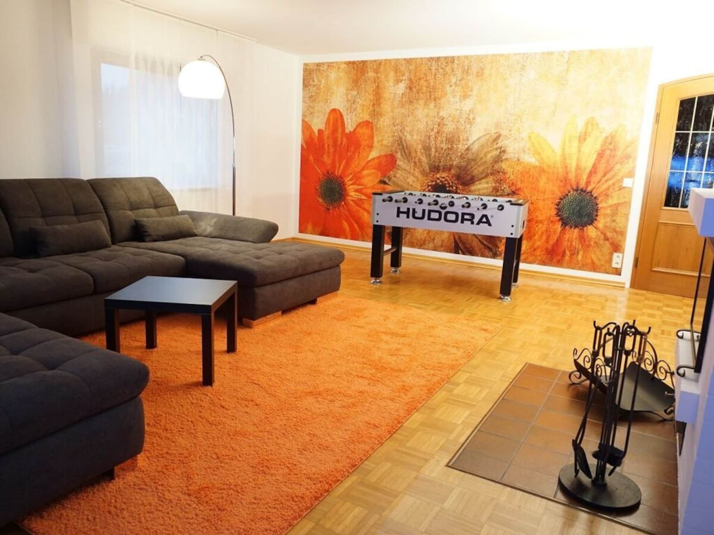 Een woonkamer met een hoekbank, een salontafel, een tafelvoetbaltafel en een groot bloemig kunstwerk aan de muur.