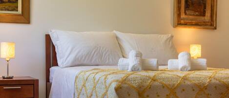 A neatly made bed with white linens and a yellow patterned bedspread, flanked by two bedside lamps and a wooden nightstand with drawers.