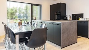 A modern kitchen with a central island, a dining area with a glass table, and a view of greenery outside. - 9 Person Holiday Home in Vaeggerlose (Vaeggerlose)