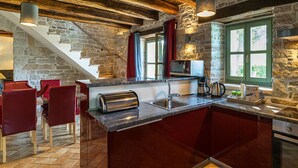 A rustic kitchen with a stone wall, wooden beams, a large island with a sink, and red chairs. - Villa Zvonar Comfortable Holiday Residence (Karojba)
