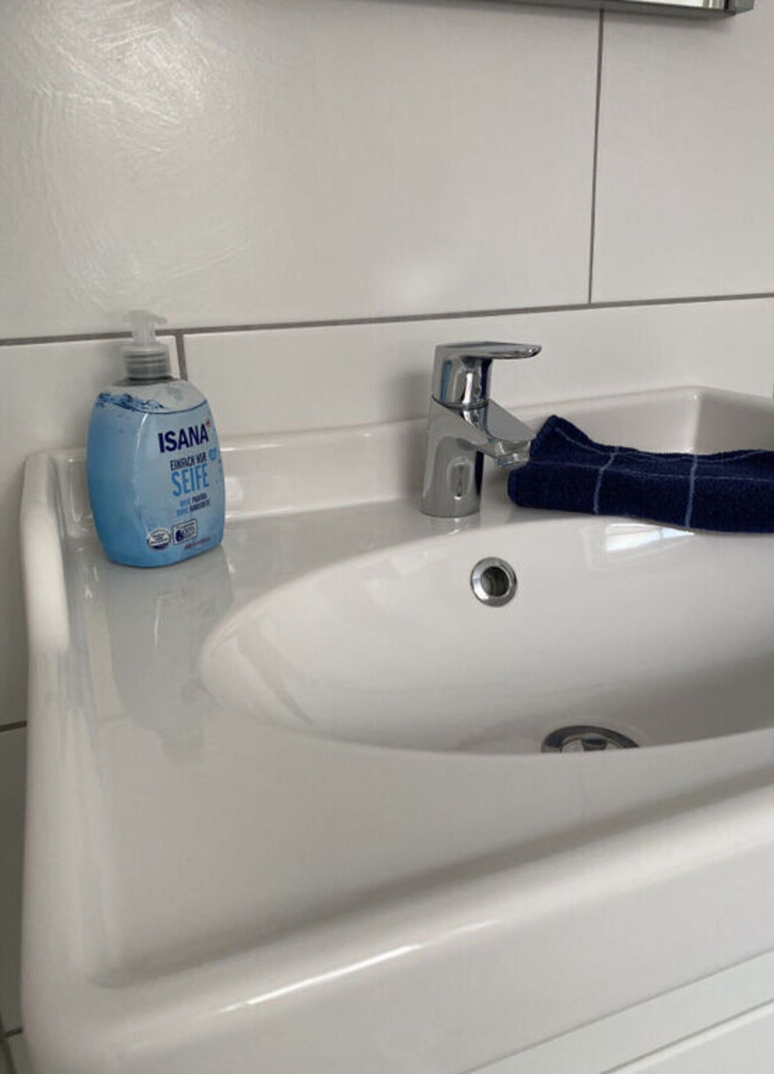 A white bathroom sink with a blue bottle of ISANA soap and a blue towel.