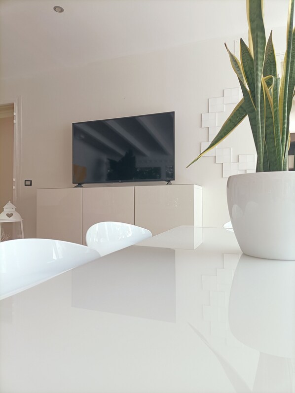 A modern living room with a flat-screen TV mounted on the wall, a white sofa, a white coffee table, and a potted plant.