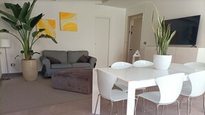 Ein modernes Wohnzimmer mit einem grauen Sofa, einem Couchtisch und einem Esstisch mit Stühlen. Zwei große Kübelpflanzen, ein flachbildfernseher an der Wand und eine Lampe auf einem Beistelltisch.