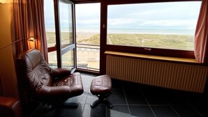 A room with a large window offering a view of the beach and dunes. There is a leather chair and a small ottoman. - Chic Urban Seaside Apartment (Nieuwpoort)