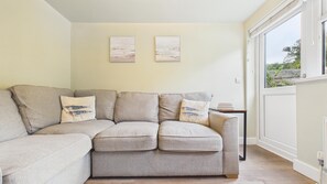 A corner living room with a beige sofa, two cushions with fish designs, and a small table with books. - Dolphin - 2 Bedroom Apartment - Saundersfoot (Saundersfoot)