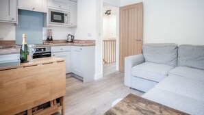 A compact kitchen with white cabinets, a microwave, and a wooden countertop. There is a grey sofa and a wooden table with a bottle and glass.