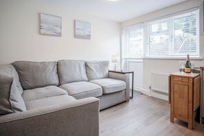 A living room with a grey sofa, two wall-mounted artworks, a side table with a lamp, and a wooden cabinet with a bottle and glass. - Dolphin - 2 Bedroom Apartment - Saundersfoot (Saundersfoot)