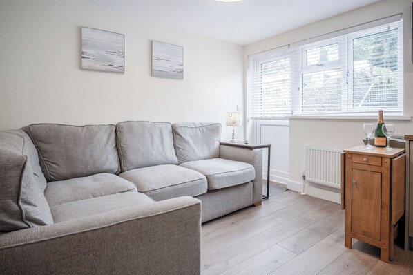 A living room with a grey sofa, two wall-mounted artworks, a side table with a lamp, and a wooden cabinet with a bottle and glass. - Dolphin - 2 Bedroom Apartment - Saundersfoot (Saundersfoot)