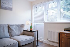 A living room with a grey sofa, a small table with a bottle and glasses, a radiator, and a window with blinds. - Dolphin - 2 Bedroom Apartment - Saundersfoot (Saundersfoot)