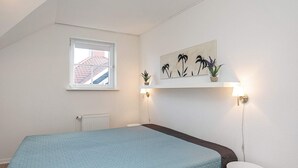 Ein Schlafzimmer mit Bett, Fenster, Heizkörper, einer an der Wand befestigten Pflanzenablage und einem Nachttisch.
