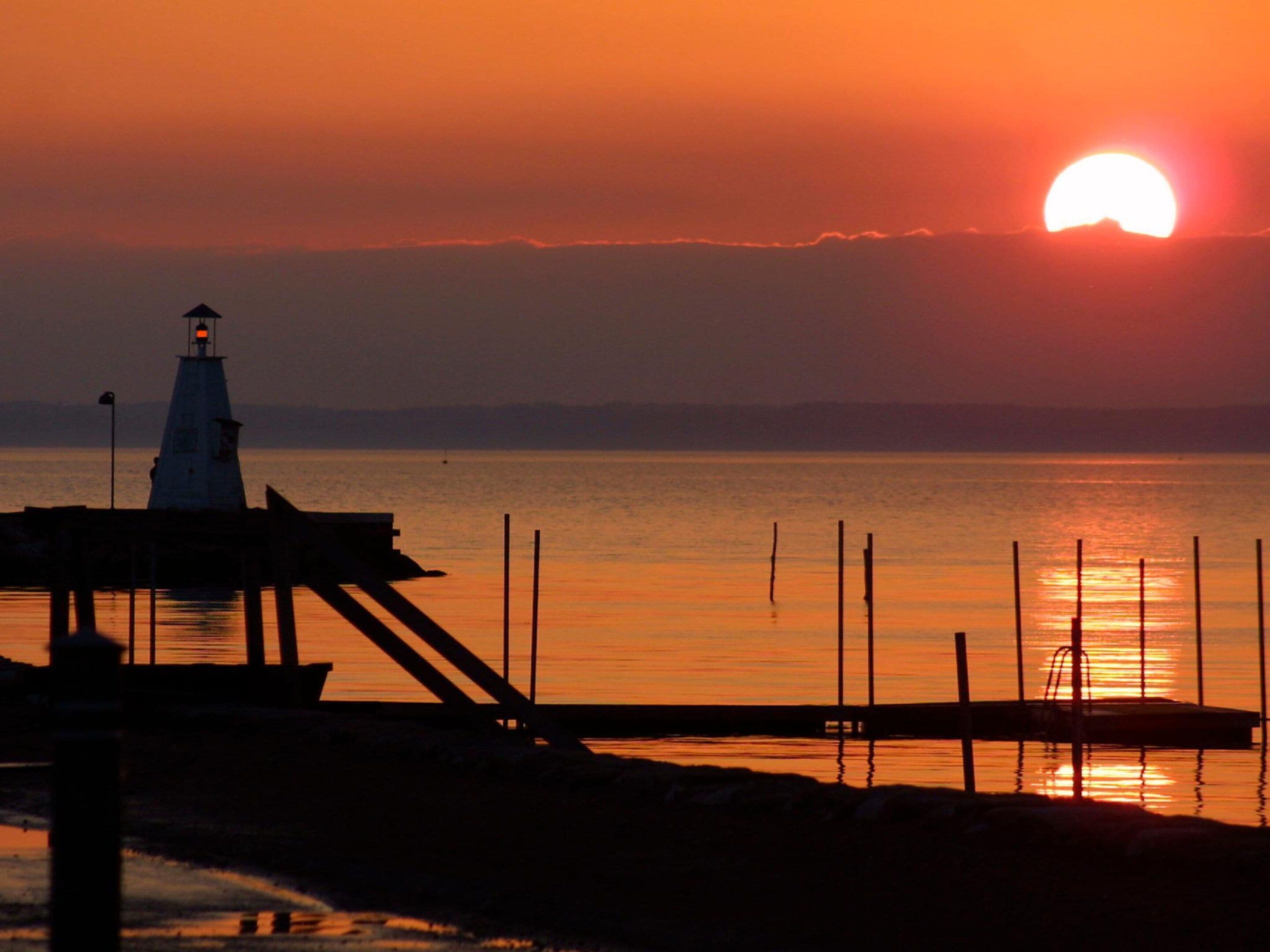 A lighthouse silhouette against a sunset, a pier extending into calm waters, and a clear horizon.