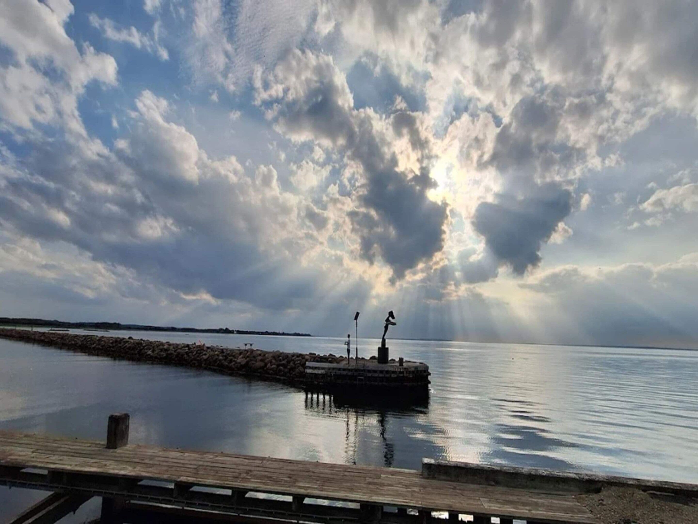 A calm body of water with a wooden dock extending into it, a rocky breakwater, and a cloudy sky.