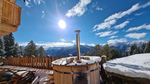A wooden sauna with a stone base, surrounded by a wooden deck and a stone wall, overlooking a snowy mountain landscape. - Hackihutte Mountain hut (Rangersdorf)