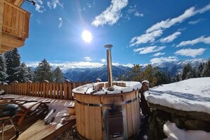A wooden sauna with a stone base, surrounded by a wooden deck and a stone wall, overlooking a snowy mountain landscape. - Hackihutte Mountain hut (Rangersdorf)