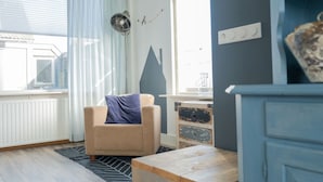 Une pièce avec un fauteuil beige, une table en bois, un radiateur et une fenêtre avec des rideaux.