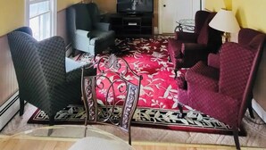 Ein Wohnzimmer mit gelben Wänden, einem roten Teppich mit Muster, zwei Sesseln, einem Sofa, einem Fernseher und einer Tür.