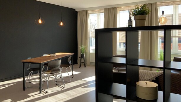 Een moderne eetruimte met een houten tafel en stoelen, een zwarte boekenkast en een kaars op een plank.
