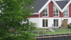 A two-story house with a red exterior and white trim, a covered patio with outdoor furniture, and a small pond in the foreground.