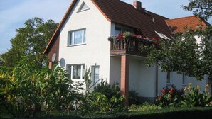 Une maison à deux étages avec un toit recouvert de tuiles rouges, un balcon orné de jardinières et un jardin où poussent diverses plantes et fleurs.