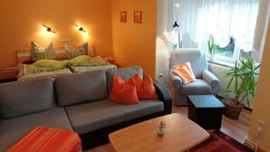 Un salon comprenant un canapé, un fauteuil et une table basse. On y trouve deux lampes fixées au mur, une fenêtre avec des rideaux légers et une plante en pot.