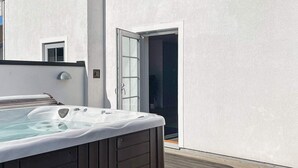 Un bain à remous sur une terrasse en bois, avec un mur blanc et une porte donnant sur un intérieur sombre.