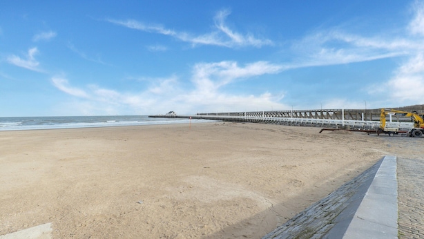 Ein Strand mit einer langen Mole, die sich ins Meer erstreckt, feinem Sand und einem gepflasterten Weg entlang der Küste.