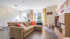 Ein modernes Wohnzimmer mit einem Ecksofa, einem kleinen Tisch, einem Bücherregal und einem Fenster mit Vorhängen.