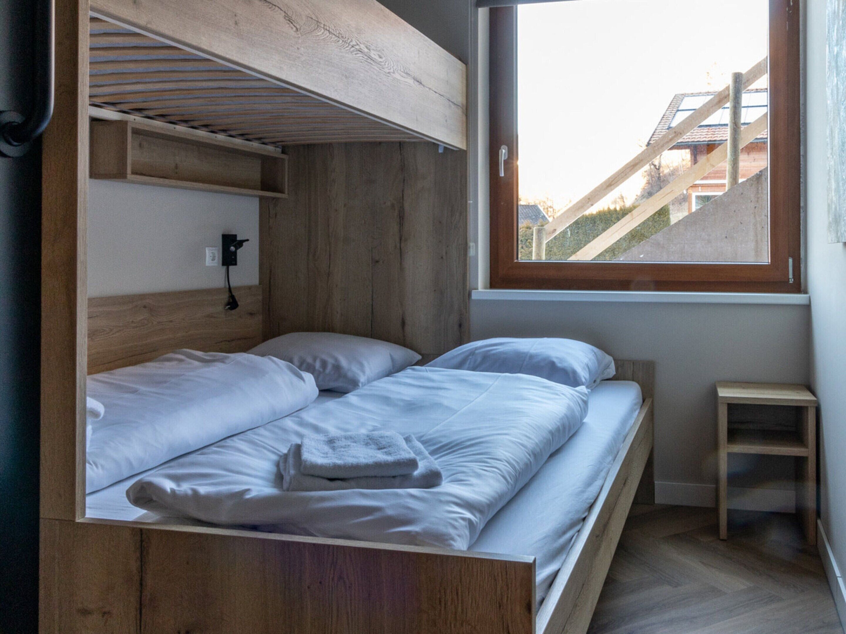 A room with a bunk bed, a small wooden stool, and a window with a view of buildings.