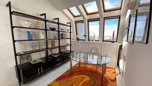 Un salon moderne avec une table en verre, des étagères noires ornées d’objets décoratifs, un tapis coloré et de grandes fenêtres offrant une vue sur la ville.