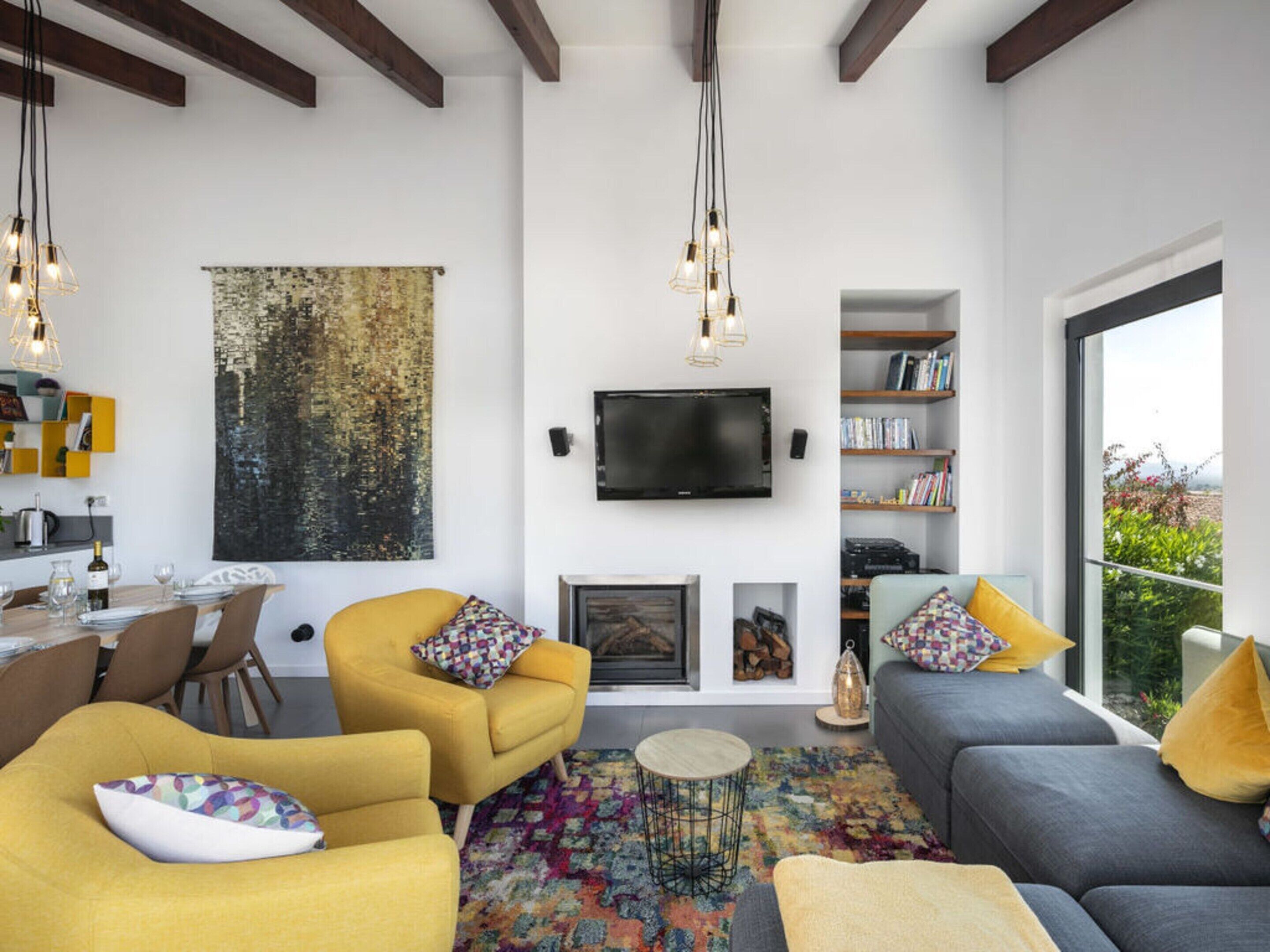 A modern living room with a fireplace, a television mounted on the wall, and a large window offering a view of greenery.