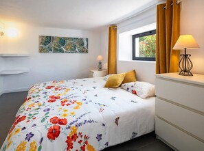 Un dormitorio ordenado con colcha estampada, una mesita de noche con lámpara, una ventana con cortinas y un cuadro colgado en la pared.