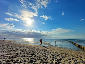 Ein Strand mit einem Steg, der sich ins Wasser erstreckt, bewölkter Himmel und einige Personen, die am Ufer entlanggehen.