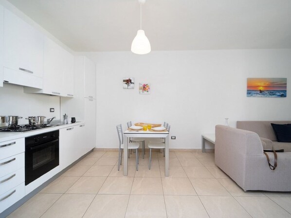 A modern kitchen with white cabinets, a dining table with chairs, and a sofa in a living area. - Apartment With Garden or Terrace Near sea (Isola di Capo Rizzuto)