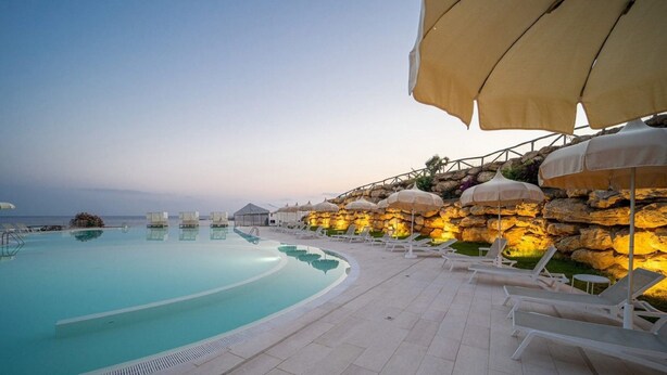 A poolside area with lounge chairs and umbrellas, a clear pool, and a rocky wall with warm lighting. - Apartment With Garden or Terrace Near sea (Isola di Capo Rizzuto)