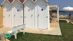 A row of white beach huts with doors, a bench, and a covered seating area with umbrellas. - Apartment With Garden or Terrace Near sea (Isola di Capo Rizzuto)