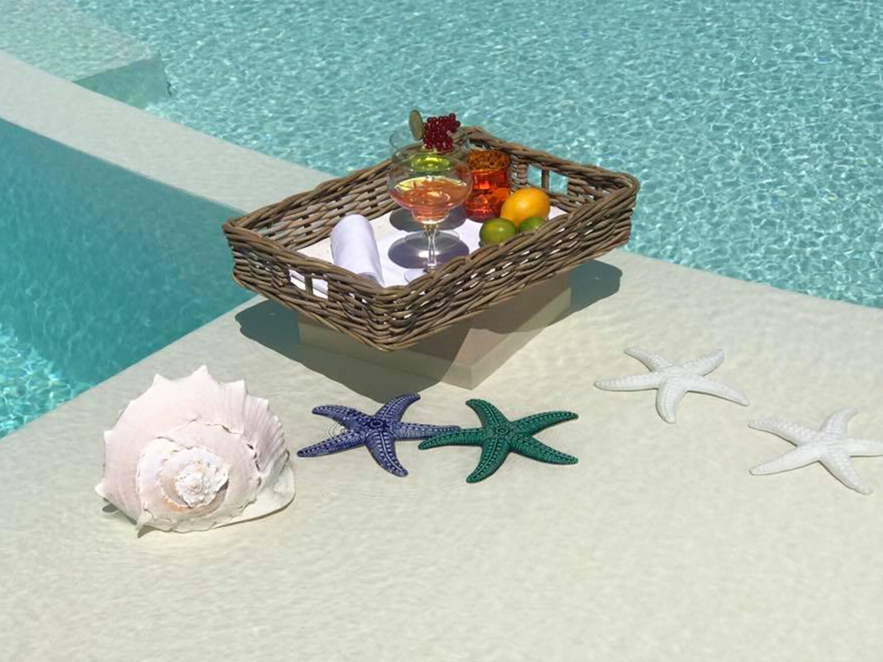 Un panier en osier contenant des fruits, un verre et une serviette, près d’une piscine décorée d’étoiles de mer et d’un coquillage.
