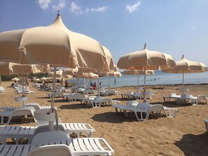A beach with rows of white lounge chairs and beige umbrellas. - Apartment With Garden or Terrace Near sea (Isola di Capo Rizzuto)