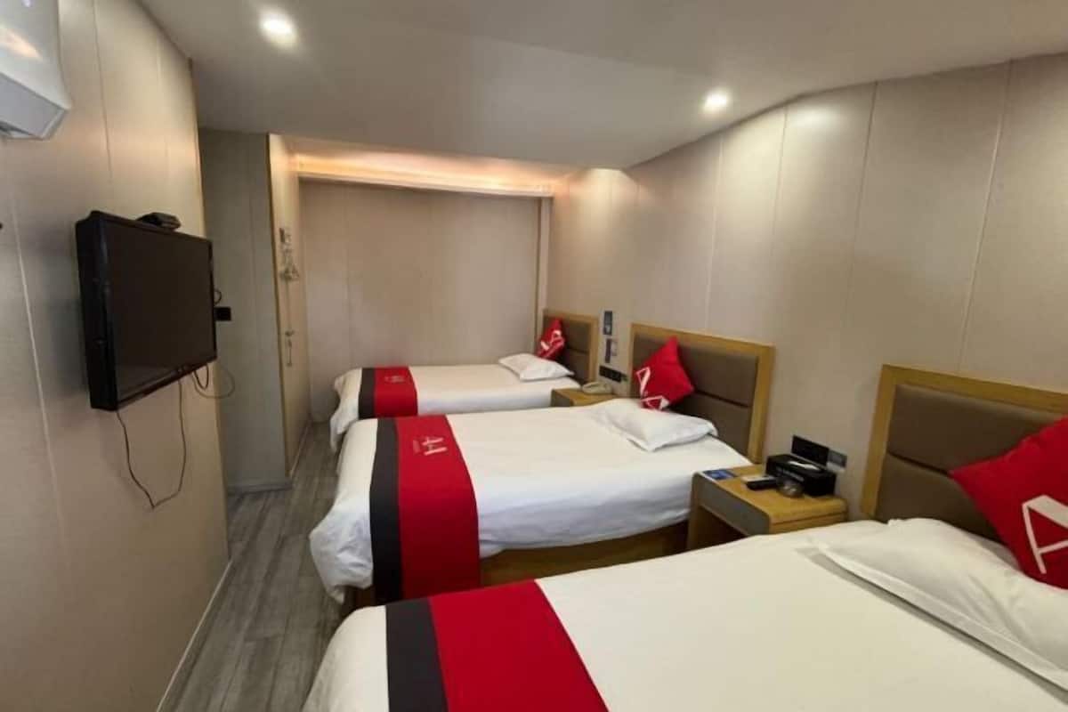 A hotel room with three beds, each with white bedding and red accents, a television mounted on the wall, and a wooden nightstand with a phone and remote control.