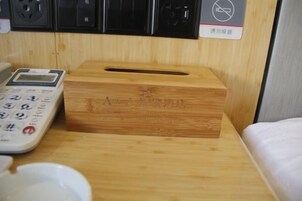 Una caja de pañuelos de madera sobre una mesita de noche, con una báscula digital y un enchufe de corriente al fondo.