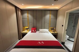 Habitación de hotel con cama, colcha roja y negra, cabina de ducha de vidrio y mesita de noche de madera con un teléfono.
