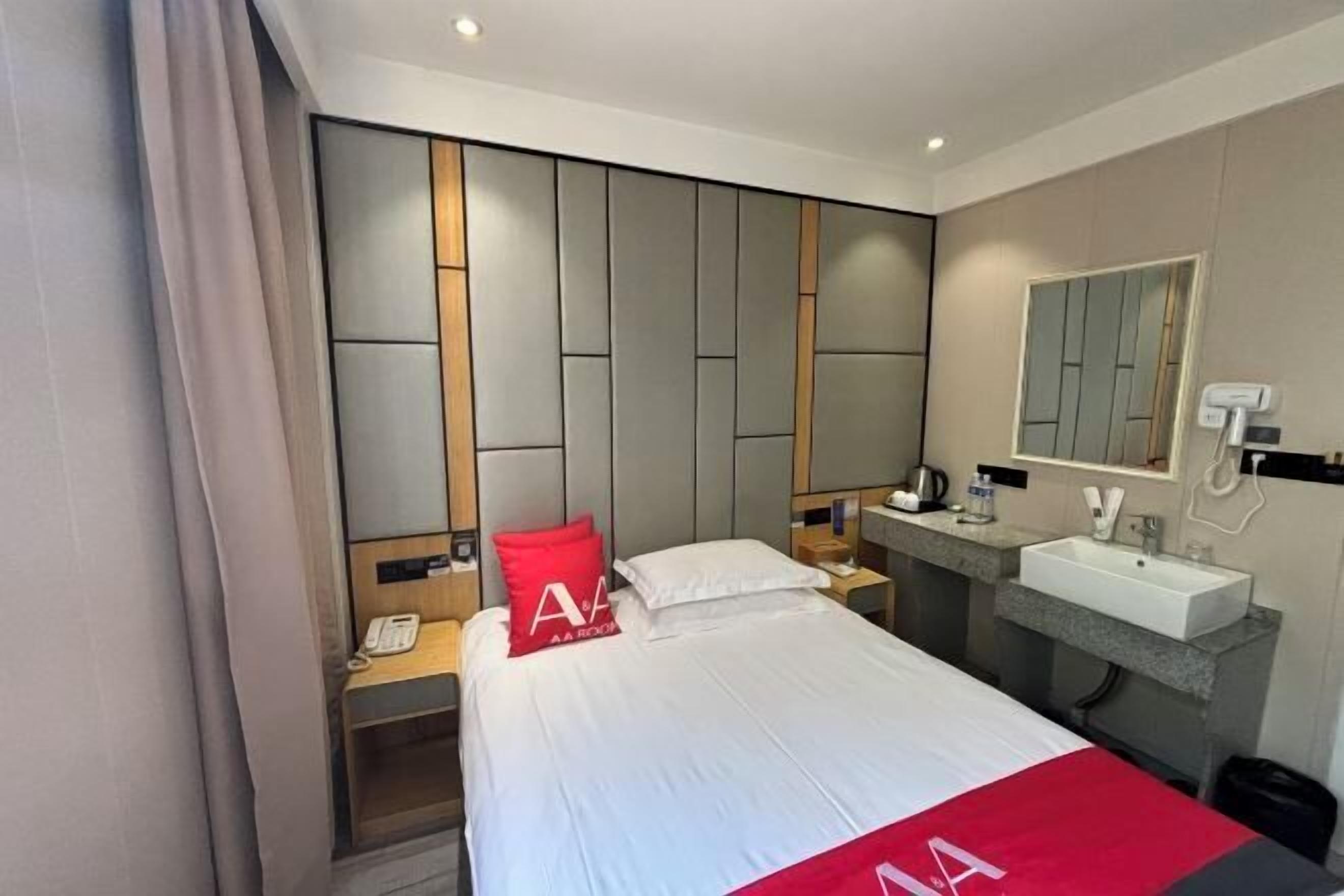 A hotel room with a bed, a bedside table with a phone, a red pillow, a nightstand, a mirror, a sink, and a shower area.
