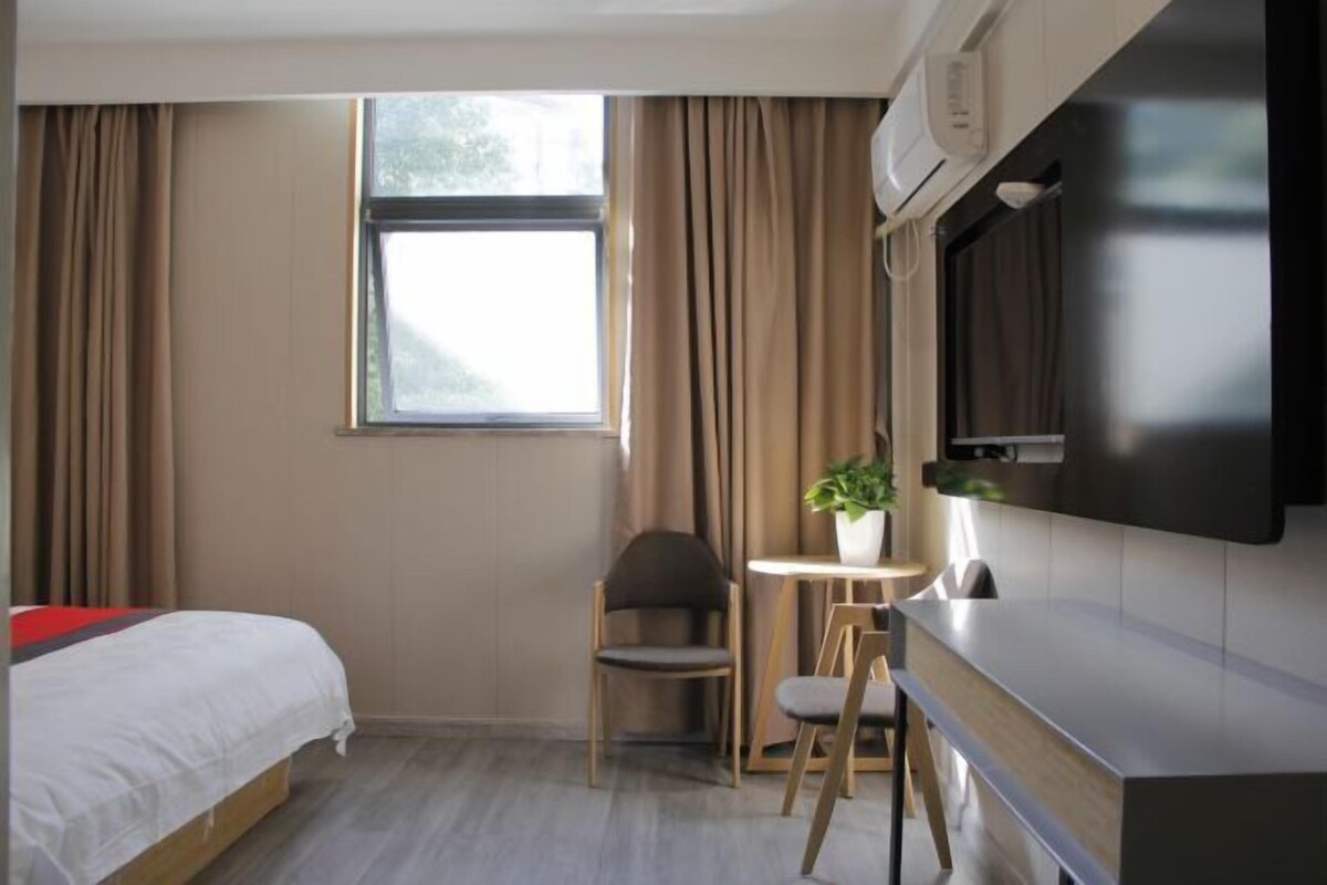 Ett hotellrum med en säng, ett skrivbord, en stol, ett litet bord med en växt och ett fönster med gardiner.